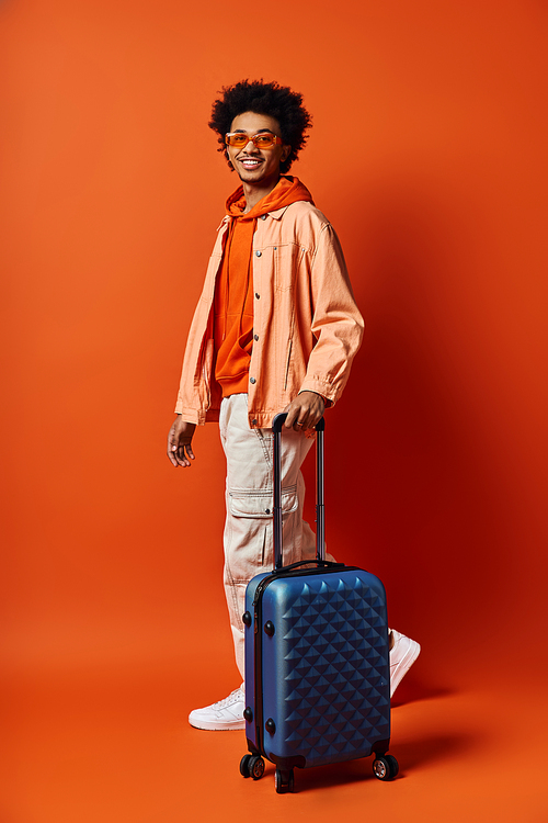 A stylish young African American man standing with a suitcase in front of an orange wall, exuding confidence and charisma.