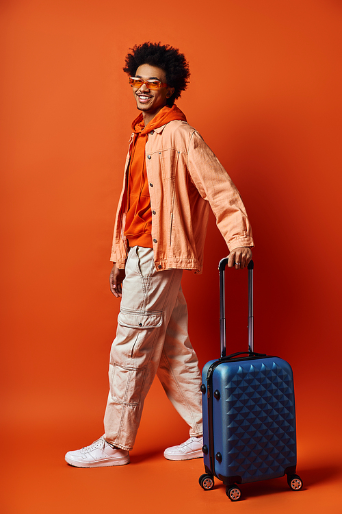 A trendy African American man with a suitcase stands confidently against a vibrant orange backdrop, exuding a sense of adventure and style.