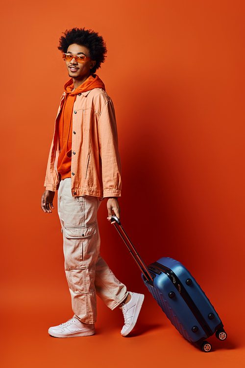 A fashionable African American man with curly hair in an orange jacket carrying a blue suitcase on an orange background.