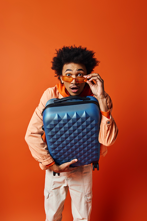 Trendy African American man with curly hair holding a stylish blue luggage on an orange background.