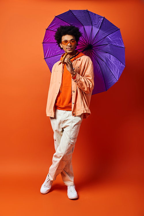 A young African American man with curly hair holds a purple umbrella against a bold orange backdrop, exuding style and personality.