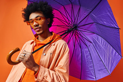 Young African American man with curly hair and sunglasses holding a purple umbrella over his head on an orange background.