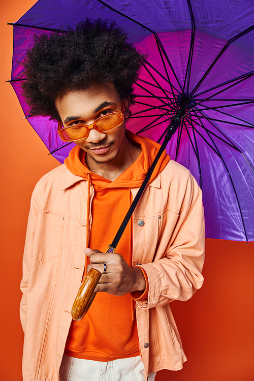 A trendy young African American man with curly hair holding a purple umbrella, exuding emotion on an orange background.