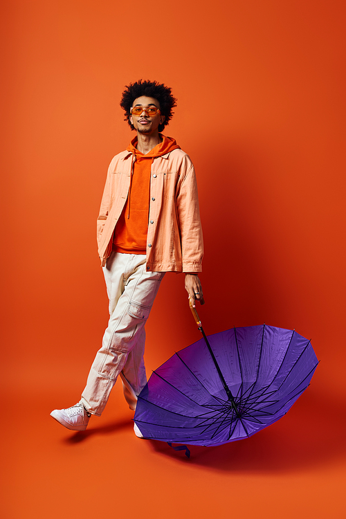 A stylish, young African American man with curly hair stands next to a purple umbrella on an orange background.