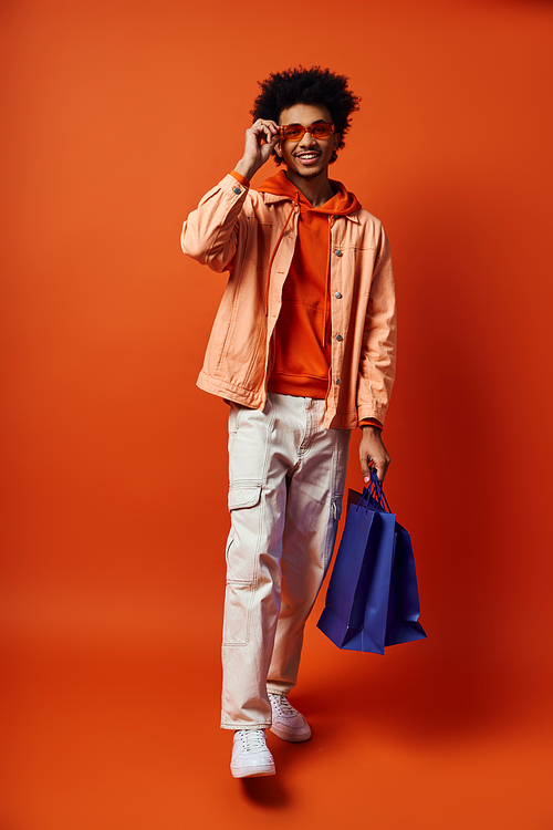 Young African American man in trendy attire, curly hair, and sunglasses, holding a blue bag while talking on a cell phone.