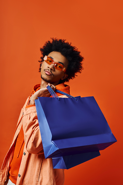 A curly African American man in trendy attire holds a blue shopping bag against an orange background, looking emotional.