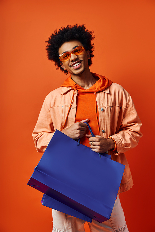 Stylish young African American man in sunglasses holding a blue shopping bag against an orange background.