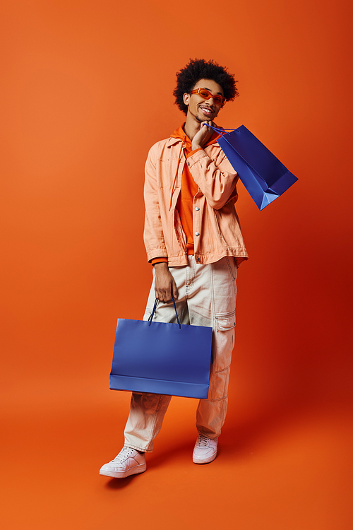 Curly African American man in trendy attire holds a blue bag while showcasing a unique, vibrant blue bag against an orange background.