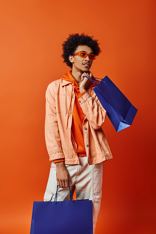 Stylish young African American man holding a blue shopping bag and looking directly at the camera on an orange background.