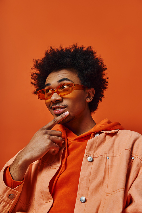 A trendy young African-American man with curly hair wearing an orange shirt and sunglasses poses against an orange background.