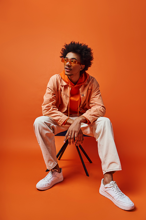 A trendy young African American man in stylish attire and sunglasses sitting on a chair