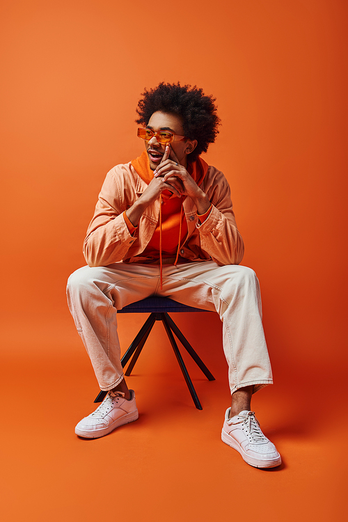 A stylish young African-American man with curly hair and sunglasses sits boldly atop a chair against an orange background.