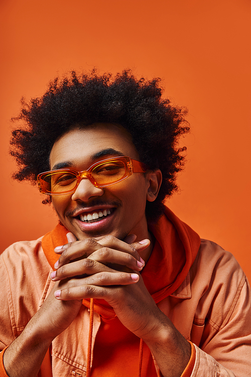 Young African American man with curly hair wearing glasses and an orange shirt against a vibrant background.