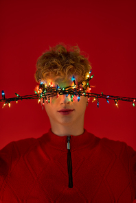 Brightly lit Christmas lights drape across a mans face, embodying the holiday spirit in a striking red setting. His joyful expression showcases his stylish seasonal outfit, perfect for celebrations.