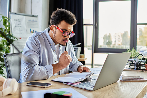 Concentrated man takes notes while working intently on a laptop in a modern office environment.