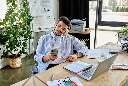 A calm, stylish man enjoys music while checking his phone in a modern office.