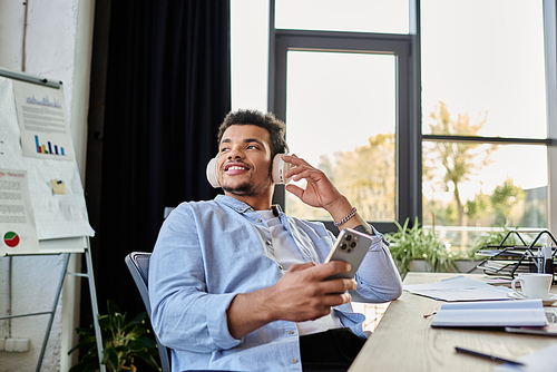 A stylish man listens to music with headphones while working at a wooden desk.