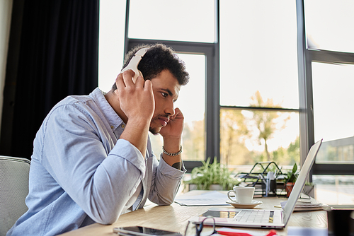 A handsome man listens intently while working on his laptop in a stylish office.