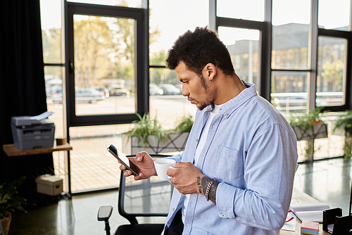 Smartly dressed man in a bright workspace enjoys coffee while engaging with his smartphone.