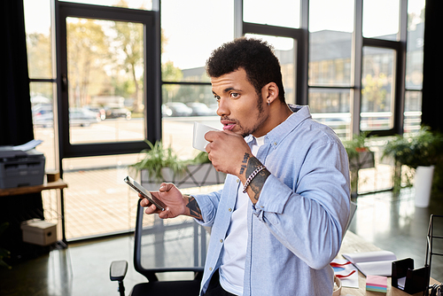 Handsome individual sipping coffee and browsing his smartphone in a stylish workspace.