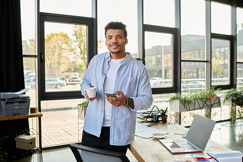 A stylish man stands in a bright office space, sipping coffee and using his smartphone.