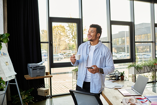 Charming man stands in a sunlit workspace, sipping coffee and engaging with his smartphone.