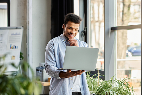 Confident young man smiles while working on laptop amidst a stylish, bright workspace with plants.