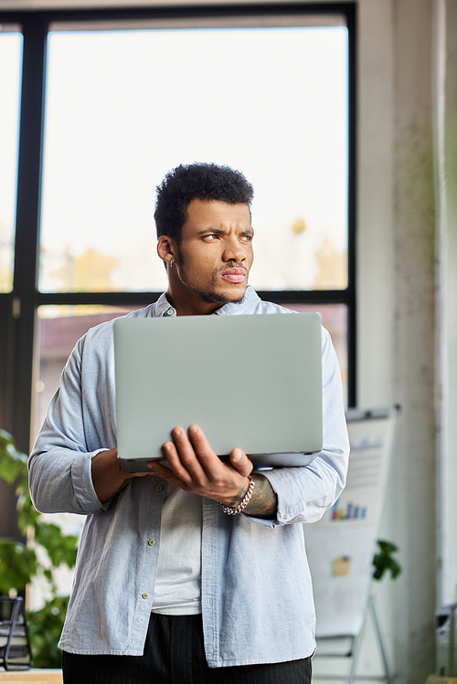 Man in a casual outfit stands holding a laptop, lost in thought amid a bright office setting.