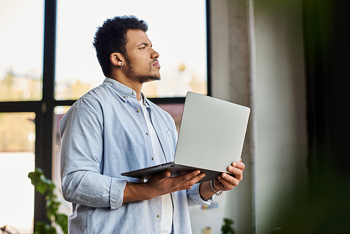 A handsome man holds a laptop while deep in thought in a sunlit space with plants.