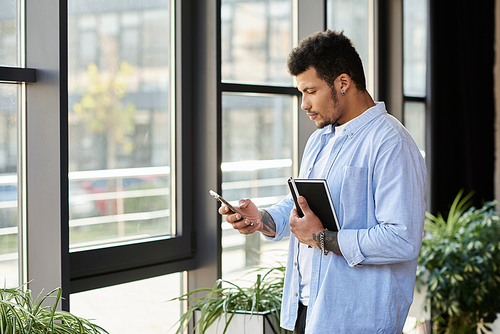 A stylish man stands by large windows, engaging with his smartphone and holding a notebook.