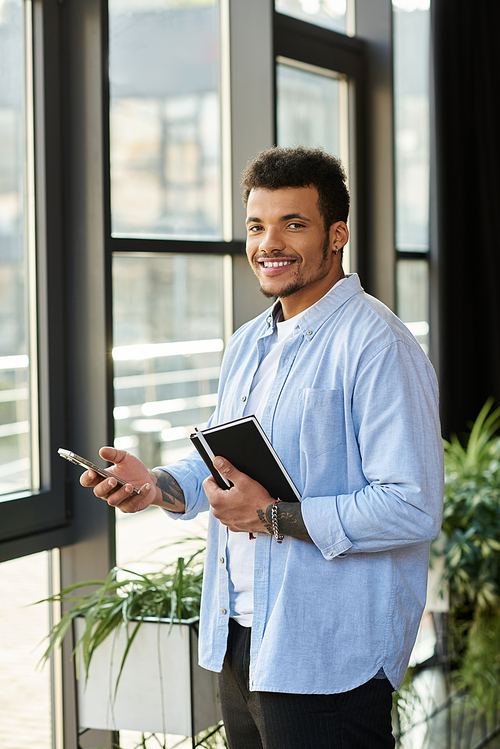 A confident man stands by large windows, smiling while holding a phone and notebook.