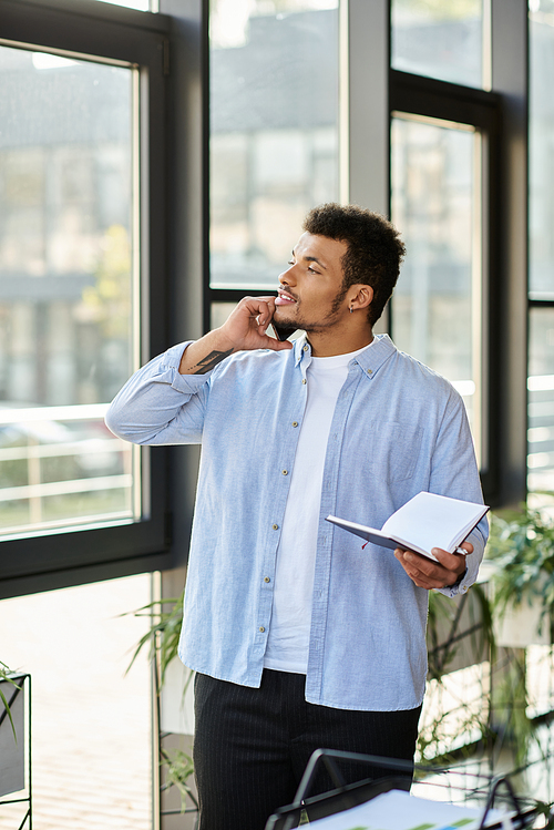 Thoughtful man holding a notebook stands by large windows in a contemporary workspace