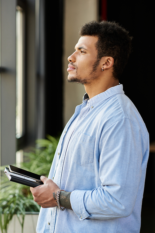 A handsome man stands near a window, contemplating with a notebook in hand, lost in thought.