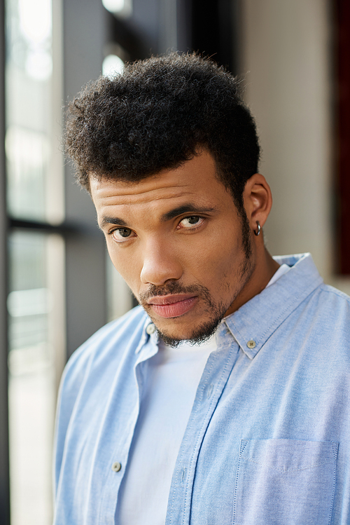 A confident young man stands by bright windows, lost in thought with an intense gaze.