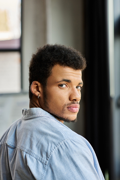 A stylish man with curly hair looks back with a contemplative expression in a bright room.