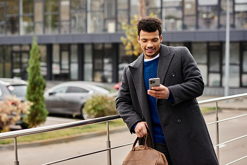 A stylish man with a bag smiles as he checks his smartphone outdoors in a cityscape.