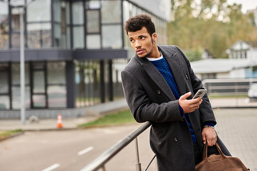 Stylish man stands in an urban environment, absorbed in his phone in the afternoon light.