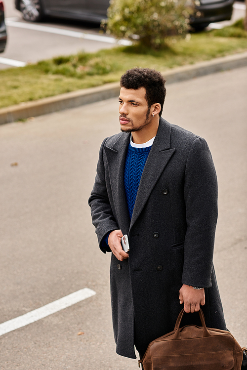 A handsome man with a confident expression strolls through a quiet parking lot holding a bag.