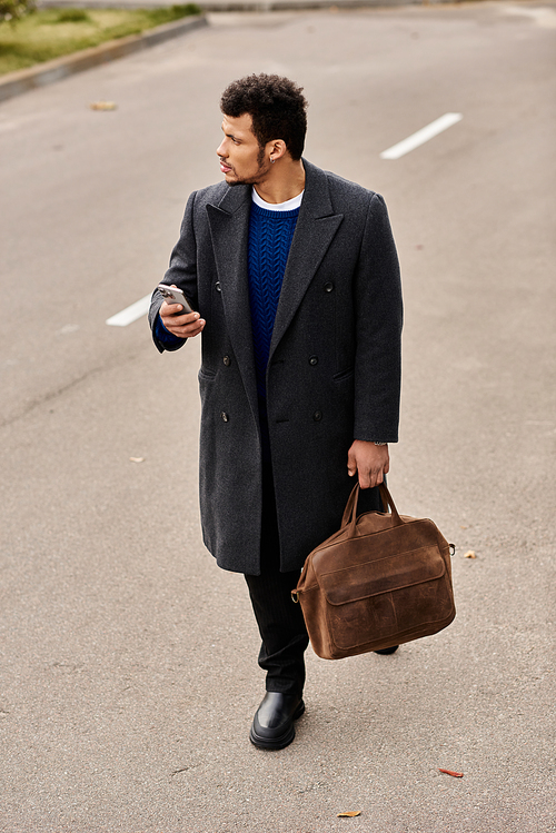 A fashionable man strolls down an urban road, checking his phone while carrying a leather bag.