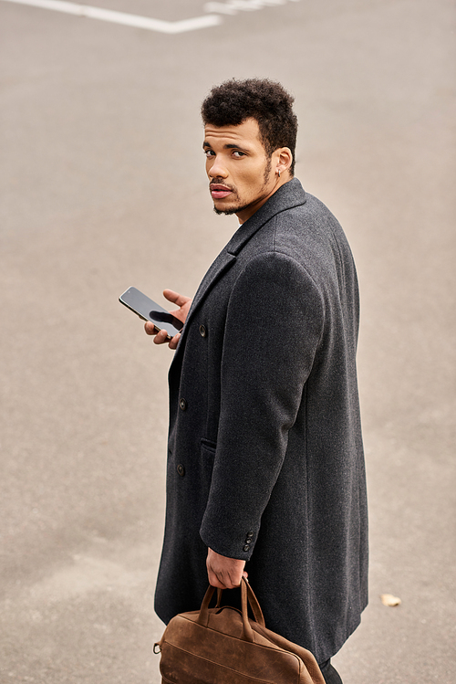 Confident man glances back while holding his phone and a brown bag, standing on the street.