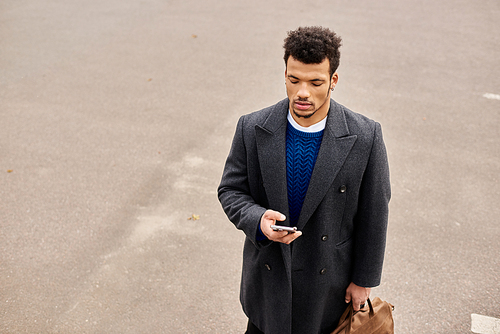 Confident man in fashionable coat engages with smartphone while strolling along quiet street.