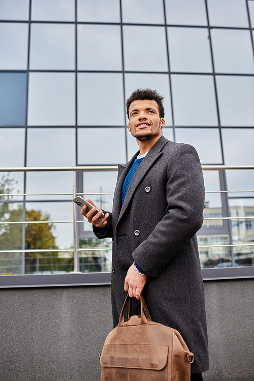 Handsome man stands outside a sleek building, smiling and holding his phone and bag.