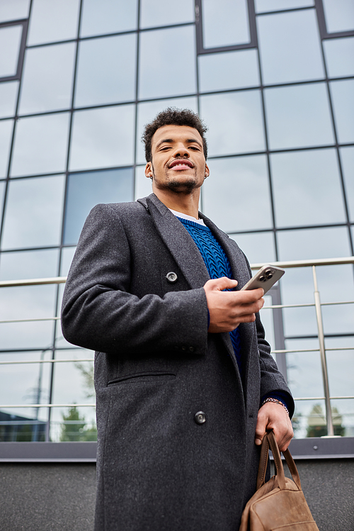 A cheerful man in a stylish coat stands outside a contemporary building while checking his phone.