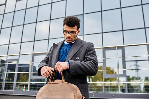 Handsome man adjusts his bag while standing near a contemporary glass building under a cloudy sky.