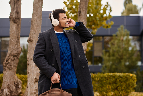 A stylish man listens to music and strolls happily among trees in a modern setting.