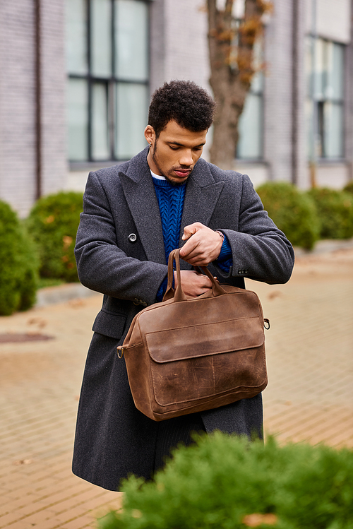 A fashionable young man retrieves items from his brown bag while walking through a city street.