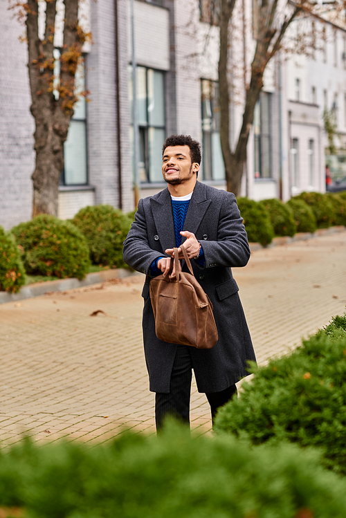 A confident man walks casually down a tree lined street holding a leather bag while smiling.