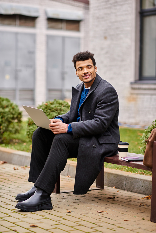 Handsome man smiles while sitting on a bench, focused on his laptop in a vibrant urban park.