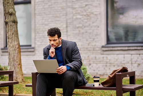 A handsome man sits on a bench, engaged in work with his laptop, enjoying natures tranquility.