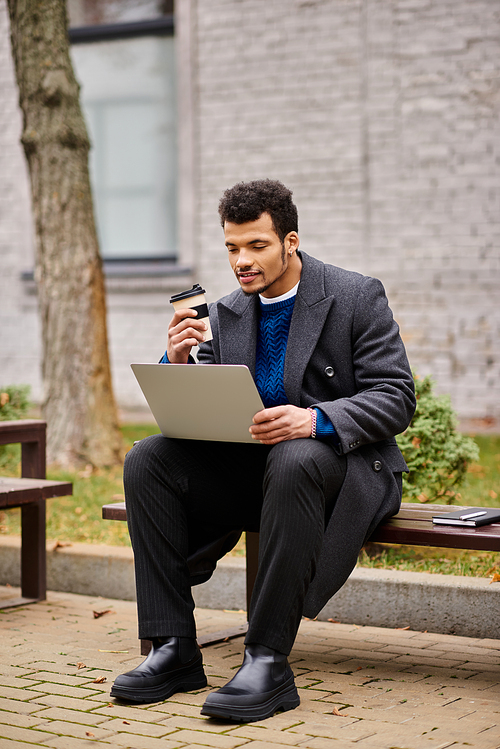 A stylish young man sips coffee and focuses on his laptop in a serene outdoor setting.